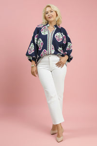 Woman wearing a navy blouse with floral patterns and white pants on a light pink background.